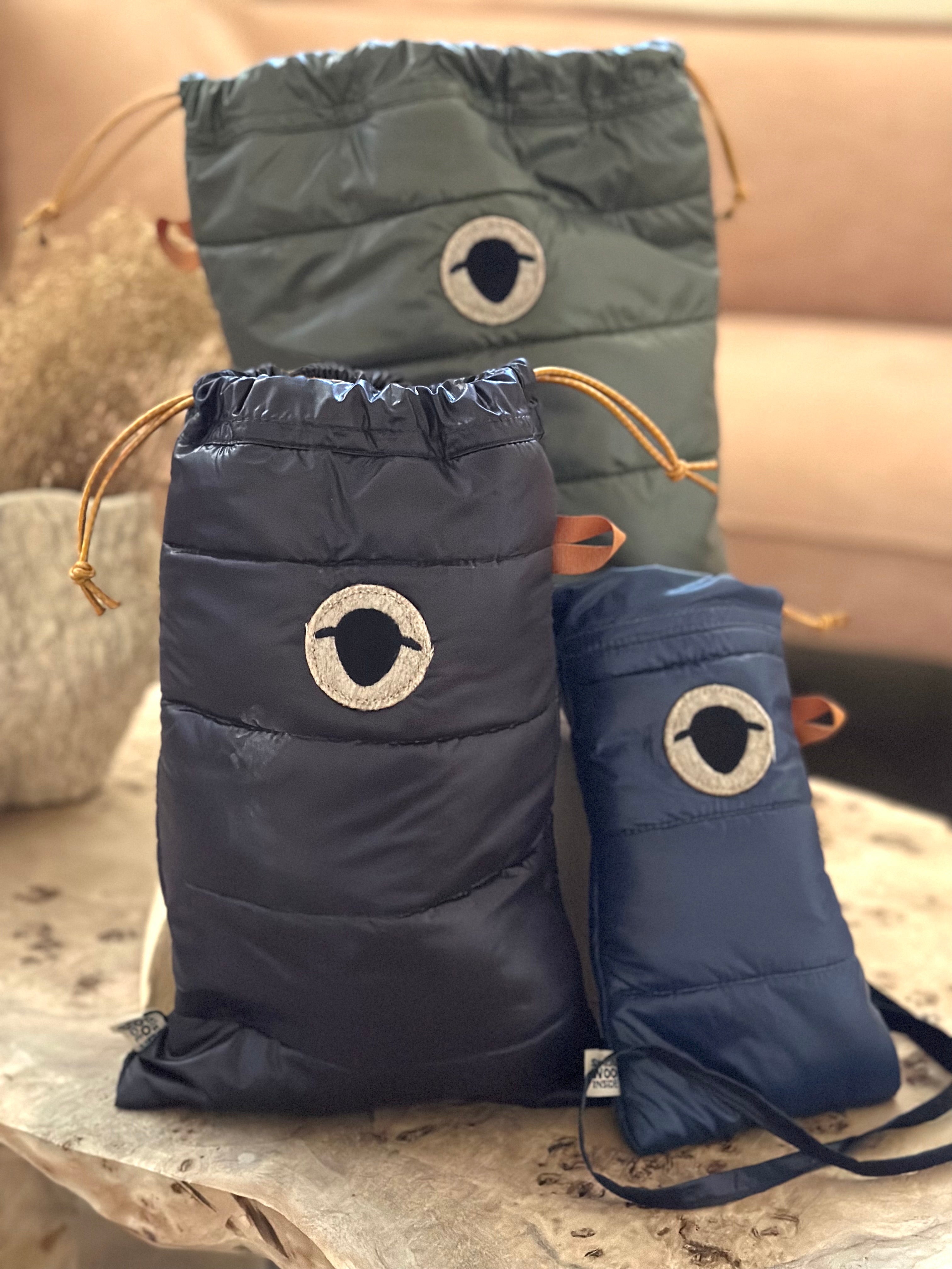 Tre funktionella påsar från Wool Rebel, uppställda mot en inomhusbakgrund. Från vänster ses en stor påse i mörkgrått, en mellanstor i svart och en mindre påse i marinblått. Varje påse har Wool Rebels logotyp och är försedda med praktiska bruna dragskoband. Den minsta påsen har även en axelrem. Påsarna är designade för att skydda innehållet mot både kyla och värme tack vare ullisoleringen.