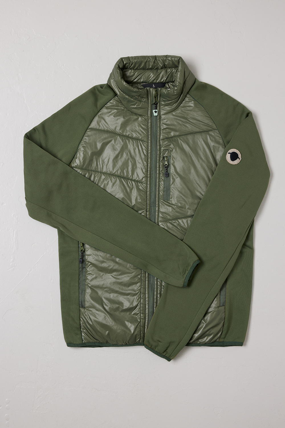 SUFFOLK Hybrid jacka, herrmodell, från Wool Rebel i färgen grön med Wool Rebel-logotyp på ena armen. Jacka är placerad på en vit bakgrund.
