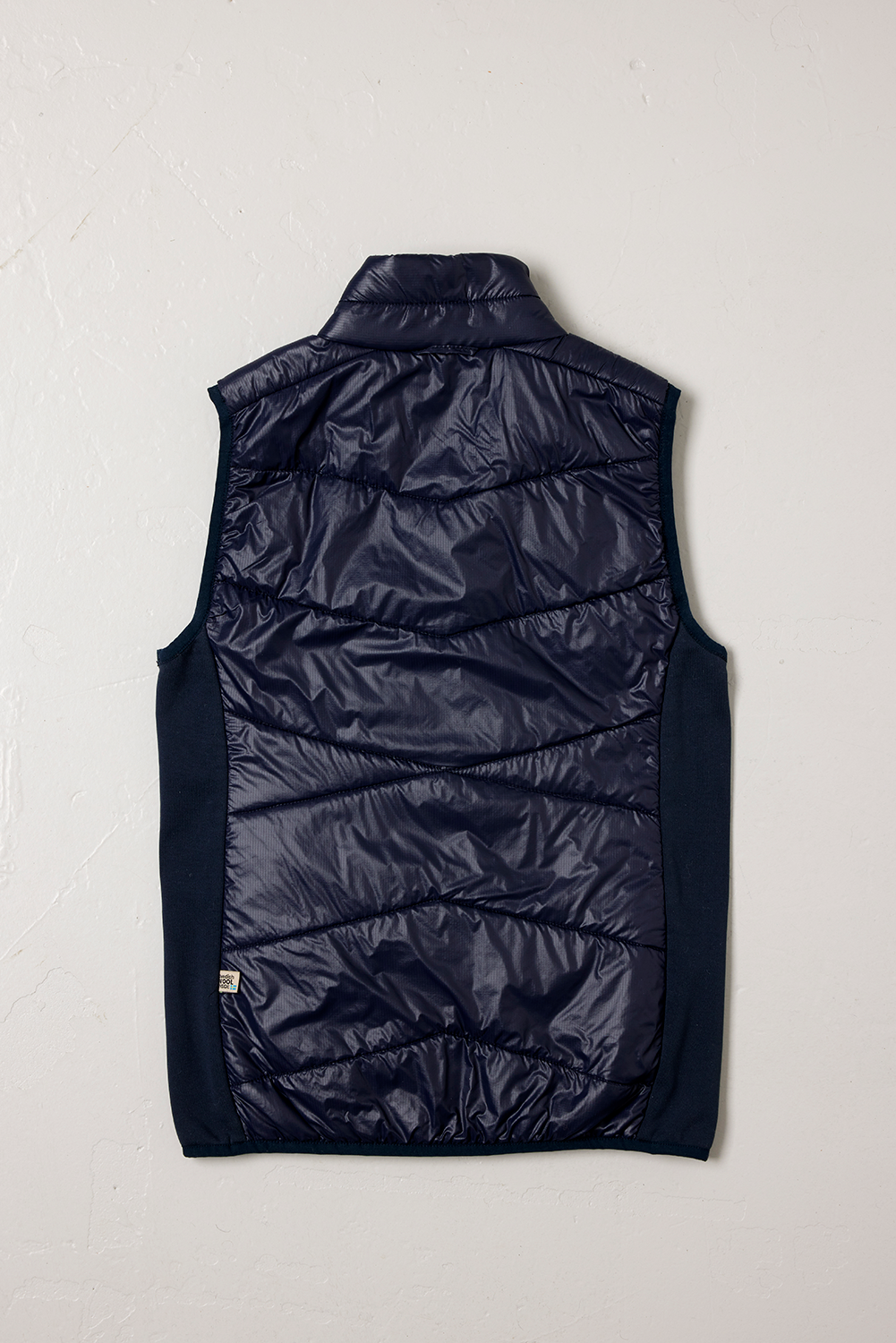 Baksidan av en Blueberry-färgad KEB Hybrid väst från Wool Rebel, med en mörkblå glansig yta och mjuka, stickade sidopaneler. Västen har även en synlig etikett från Wool Rebel nertill på höger sida, fotograferad mot en vit texturerad bakgrund.