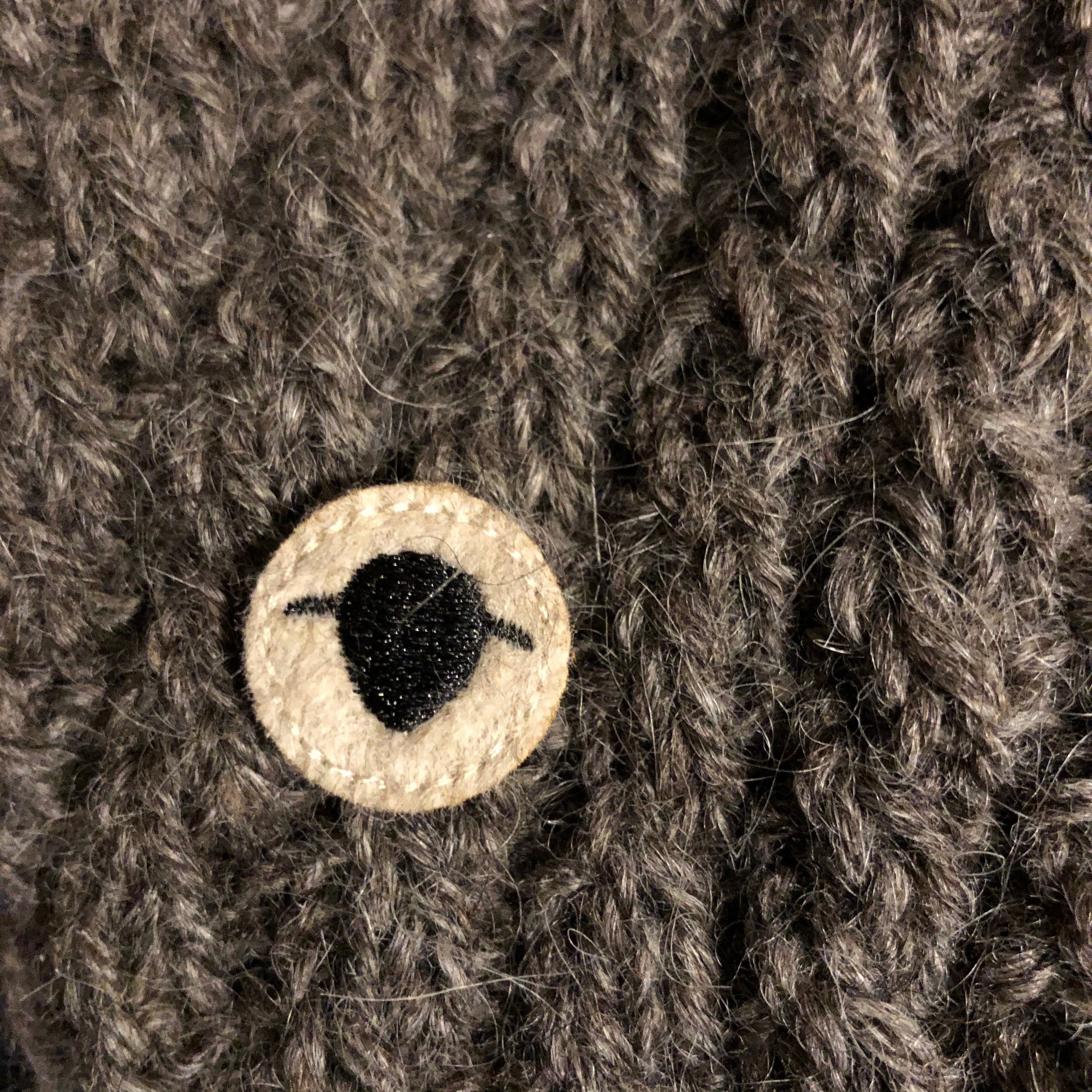 Närbild på Wool Rebel-logotypen som visar en stiliserad svart och beige fårformad symbol, fastsydd på en gråbrun ulltextur.