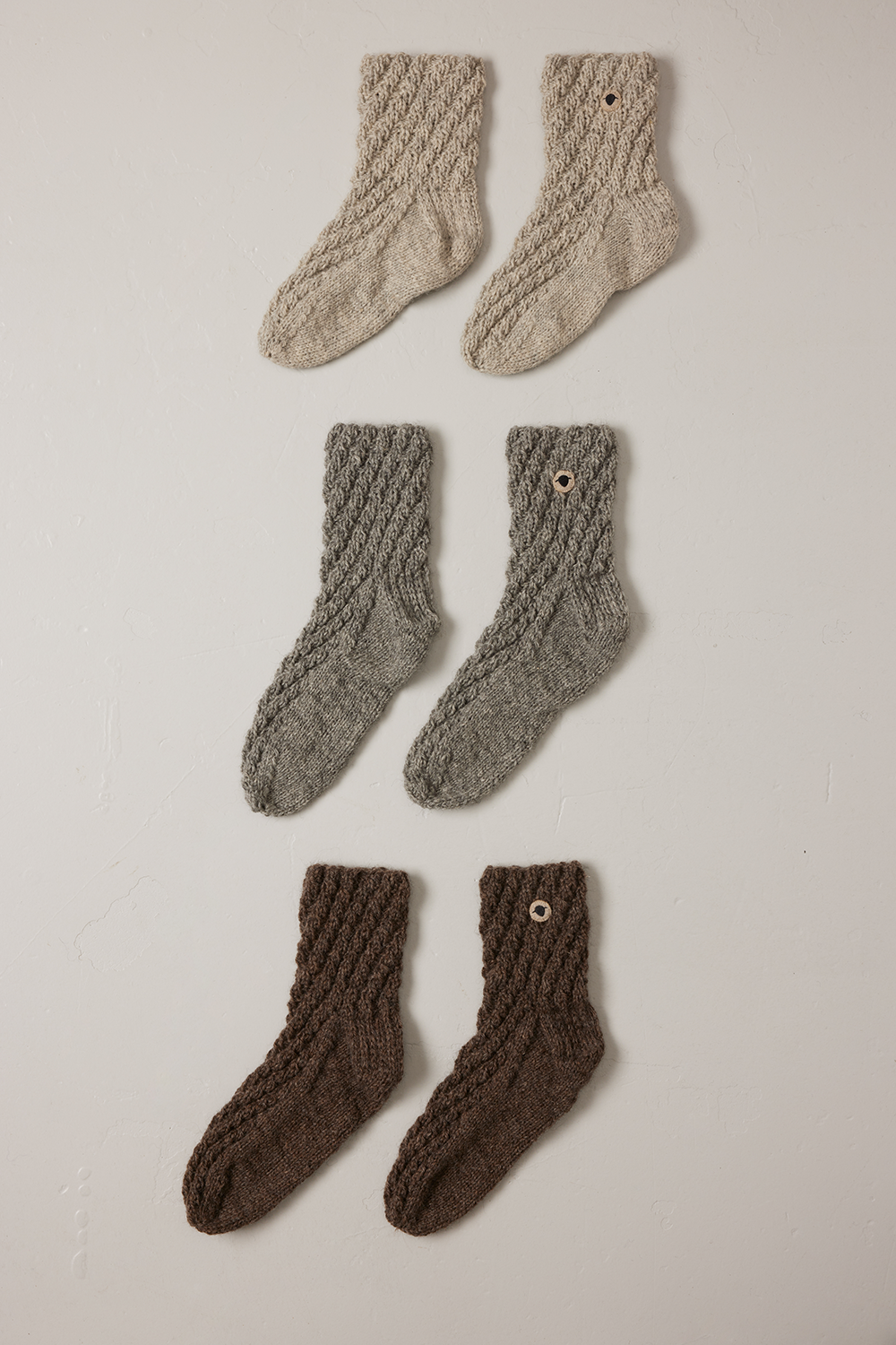 Handstickade SHETLAND-sockar från Wool Rebel, visade i tre olika nyanser av naturlig ullfärg på en vit bakgrund. Varje par har en synlig Wool Rebel-logotyp.